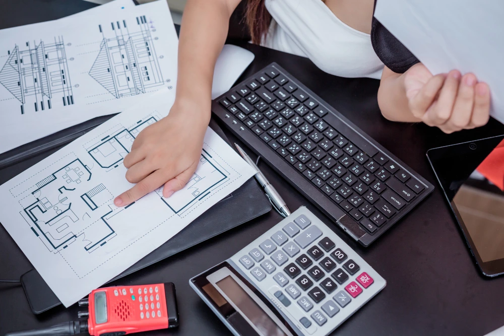 Person in white shirt reviewing detailed architectural floor plans and blueprints on dark desk, with calculator, laptop keyboard, and smartphone visible in workspace - Estimation & Takeoff Services for building Warehouses