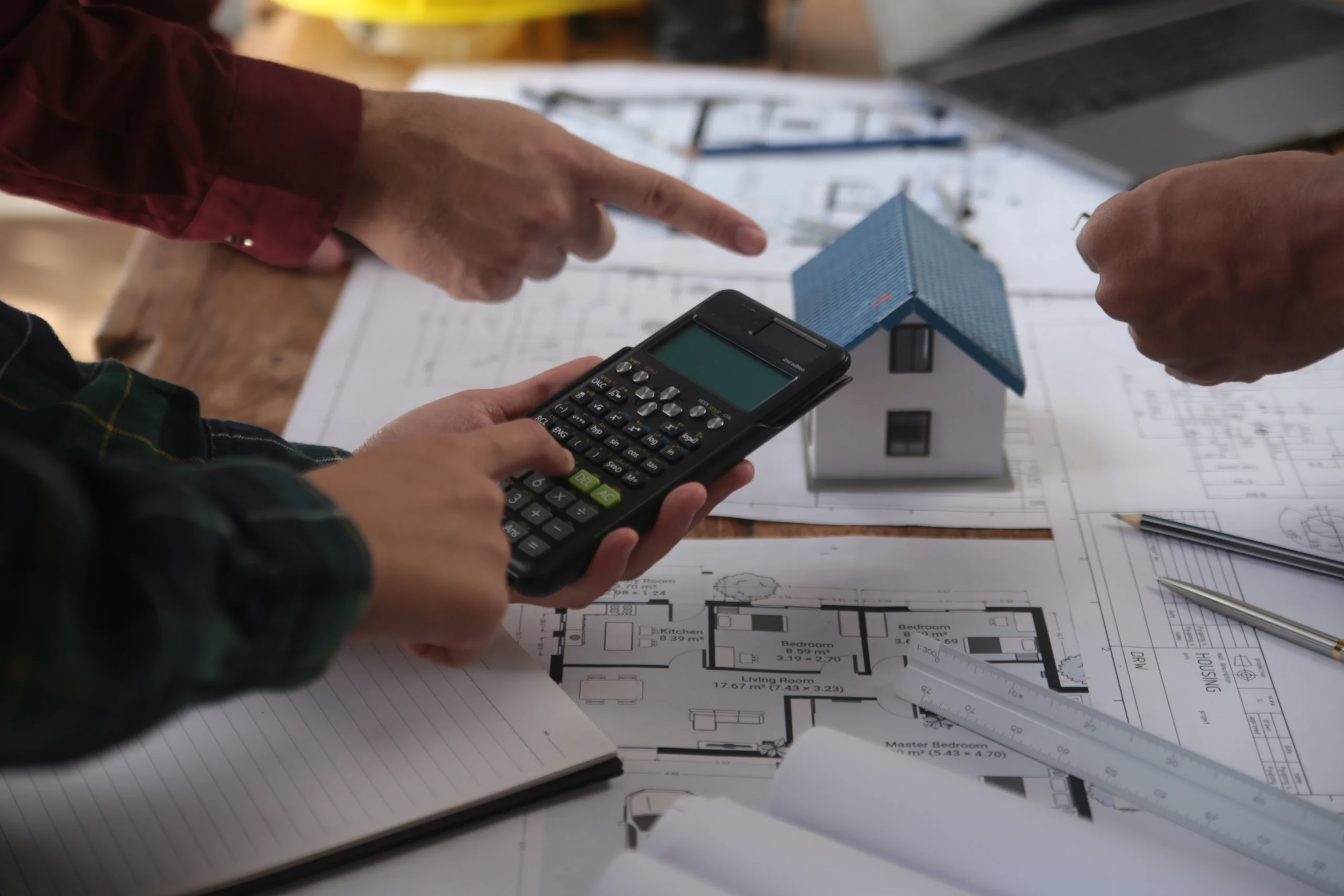 Two people collaborating over house plans and blueprints, with one person holding a calculator and a small house model visible on the table