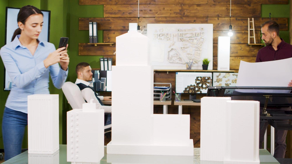 Architects in modern office examining white architectural models on table, with woman taking photo on phone while two men work at desks with technical drawings- Restaurant Renovation Estimation Builds Profit
