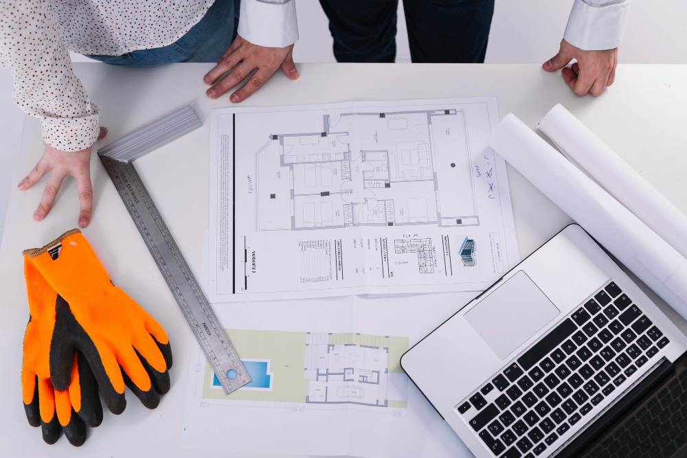 Two professionals reviewing architectural plans and blueprints on a white table, with a laptop, a ruler, and orange safety gloves nearby