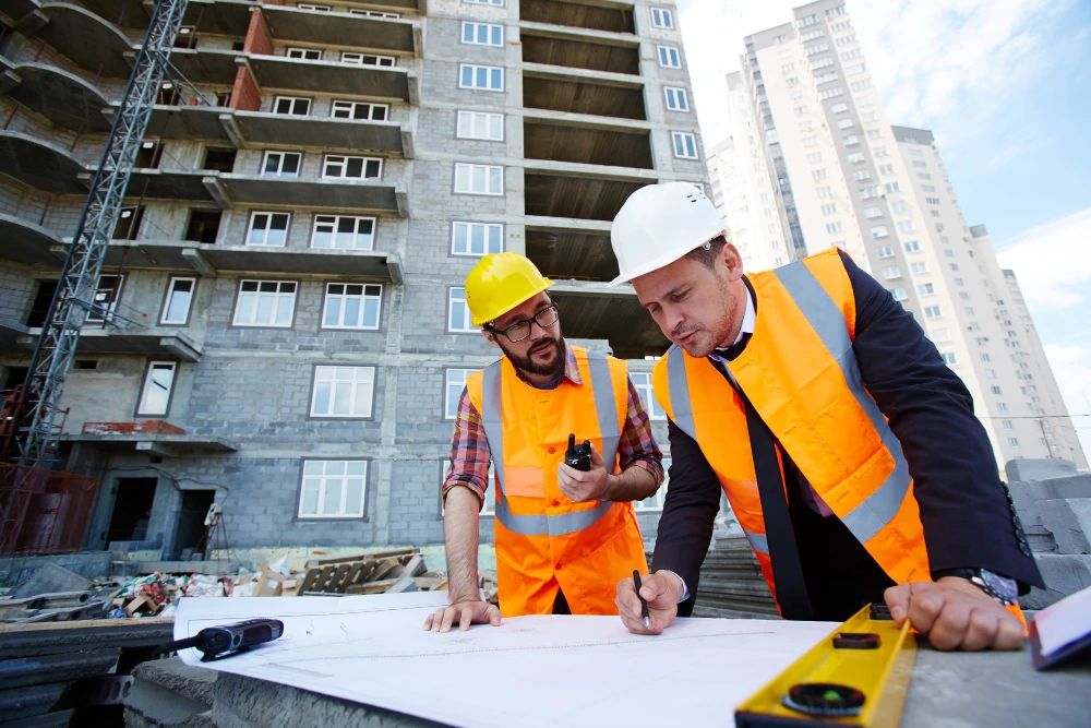 Two construction engineers wearing safety vests and helmets reviewing blueprints on a construction site with a high-rise building in the background.