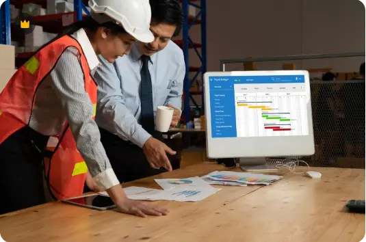 Two construction professionals in hard hats examining project plans and documents at a desk with project management software displayed on computer monitor