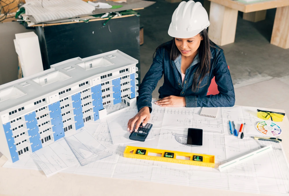 Female architect in white hard hat working with calculator and blueprints next to architectural building model in construction office - MEP Construction Estimation Services