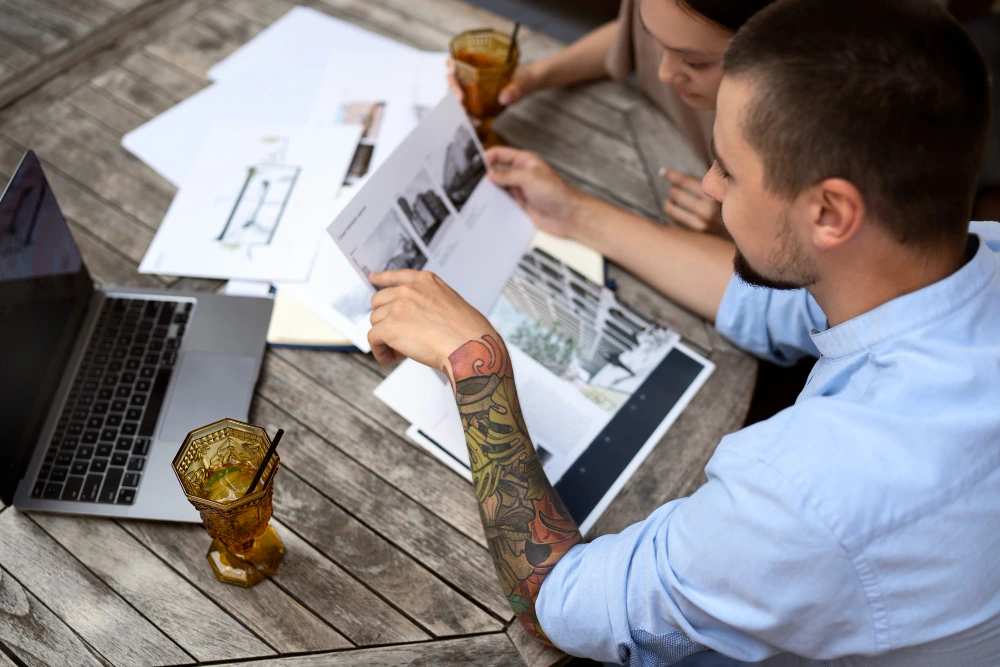 Two business professionals sitting at a wooden table reviewing architectural drawings and documents, with laptops and drinks, man wearing light blue shirt has visible tattoo on forearm- Garage Conversion Estimator