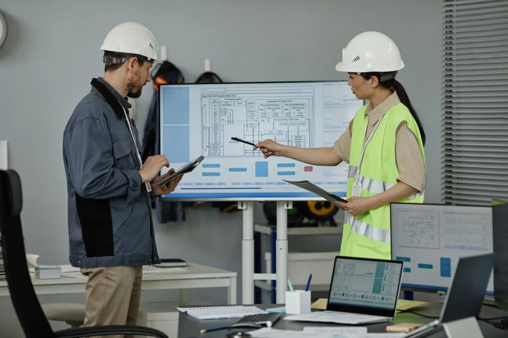 Two construction professionals in hard hats and safety vests reviewing architectural blueprints on a drafting table in an office setting, with one person pointing at technical drawings while the other holds a tablet- Concrete Estimation