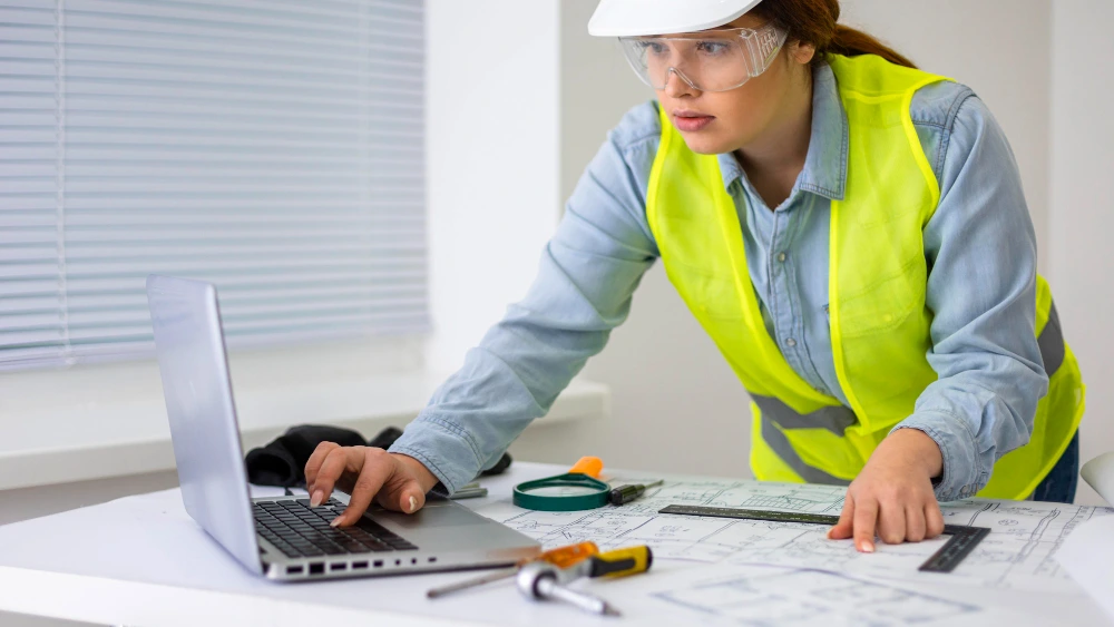 Female engineer in hard hat, safety glasses, and high-visibility vest working on laptop while reviewing technical drawings and blueprints at desk with engineering tools -