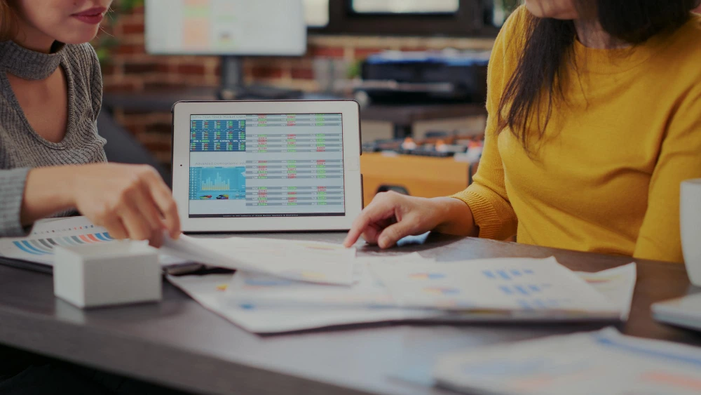 Two women analyzing financial data on a tablet displaying spreadsheets and charts, with printed financial reports and documents on the desk in an office environment