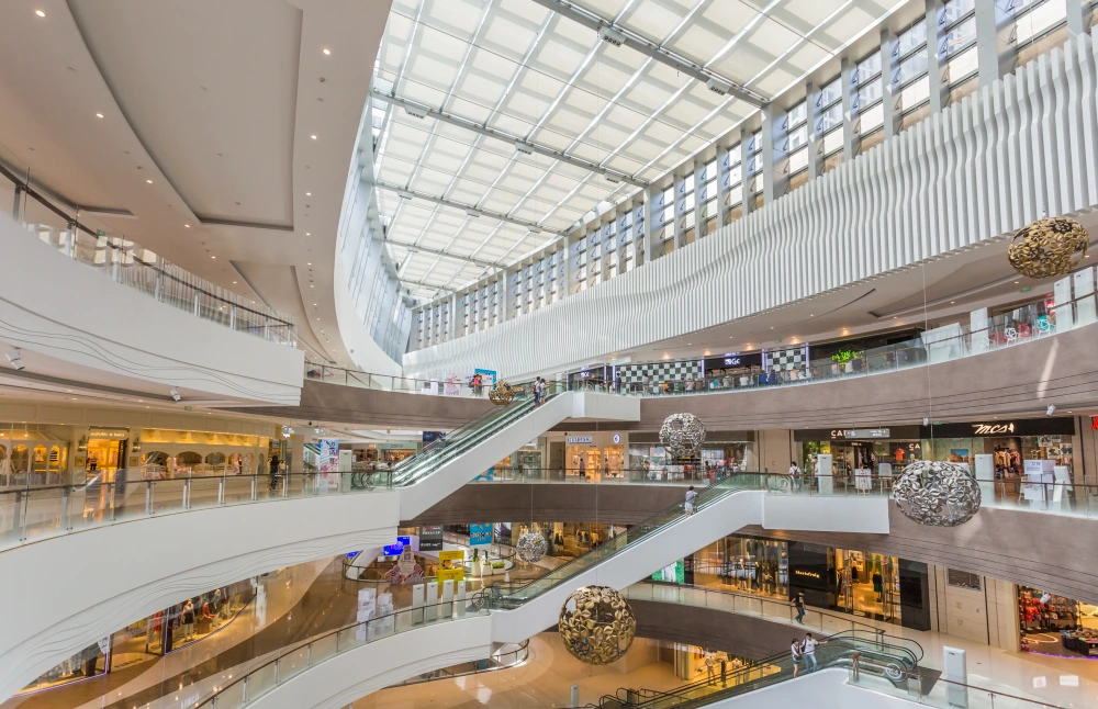 Spacious modern shopping mall interior with multiple levels, glass ceiling skylight, curved escalators, decorative spherical art installations, and retail stores on each floor