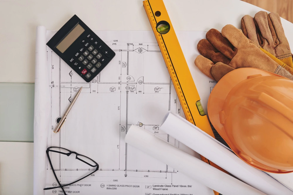 Construction workspace with architectural blueprints, orange hard hat, work gloves, calculator, spirit level, and safety glasses arranged on technical drawings  - Excel vs. estimating software