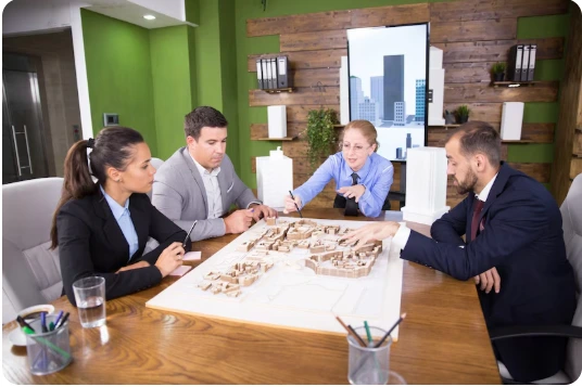 Four business professionals in suits examining architectural model and blueprints during meeting in modern conference room with green accent wall and city skyline artwork -  In-House vs Outsource Construction Estimation