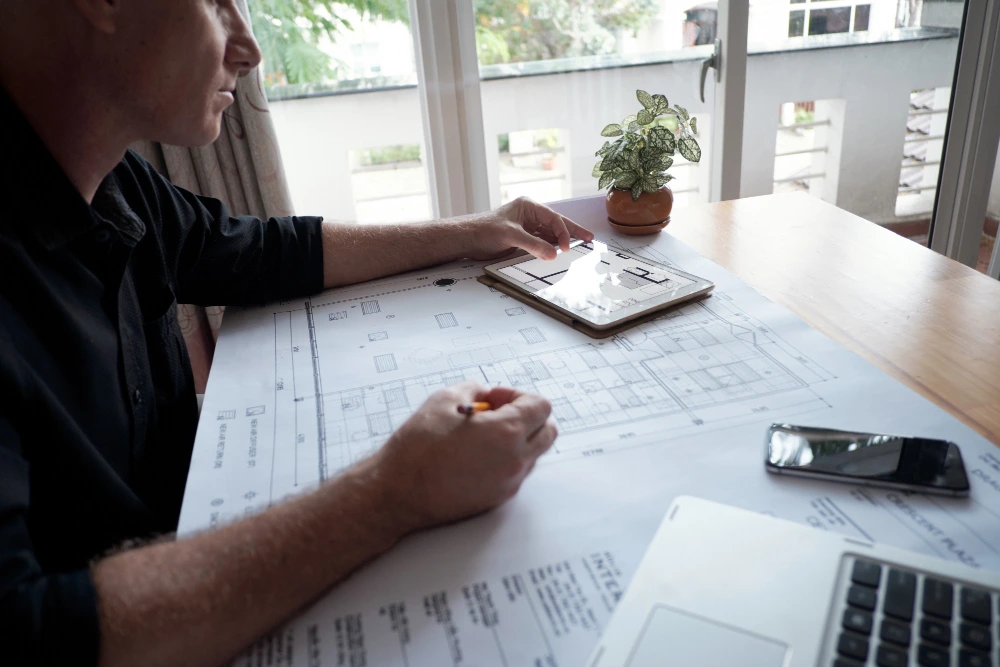 Architect's hands working on digital floor plan using tablet with stylus, surrounded by paper blueprints and calculator on desk