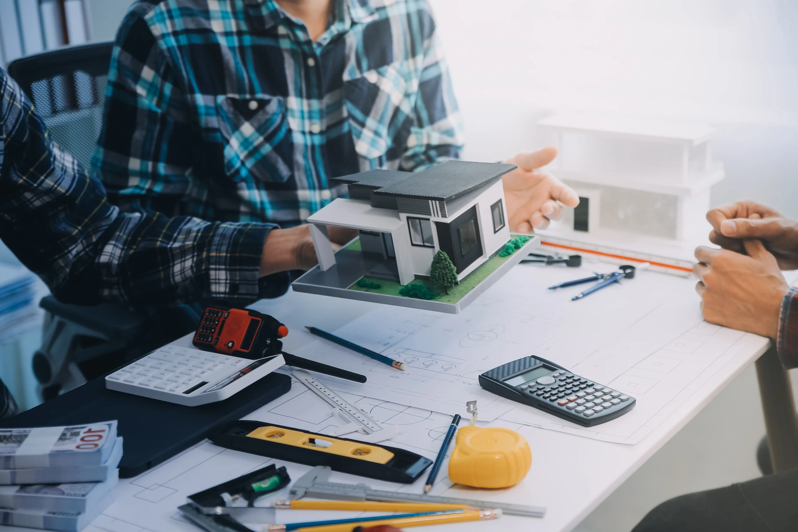 Architect or designer presenting a modern house model to clients at a desk covered with blueprints, calculators, and drafting tools