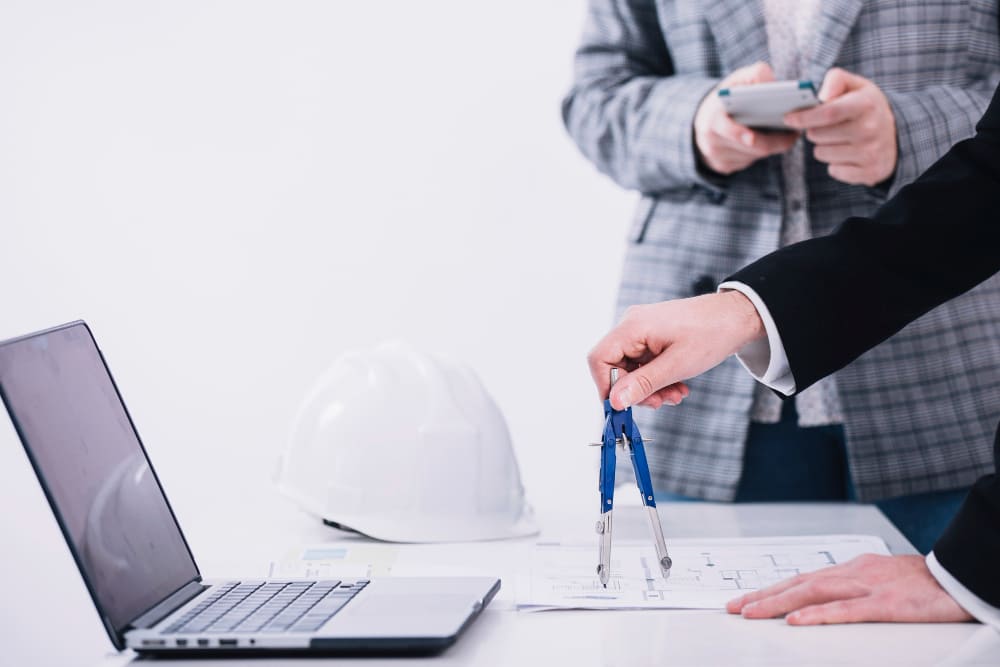 Construction professional in high-visibility safety vest using laptop computer at desk with architectural blueprints and plans. Construction project management and planning