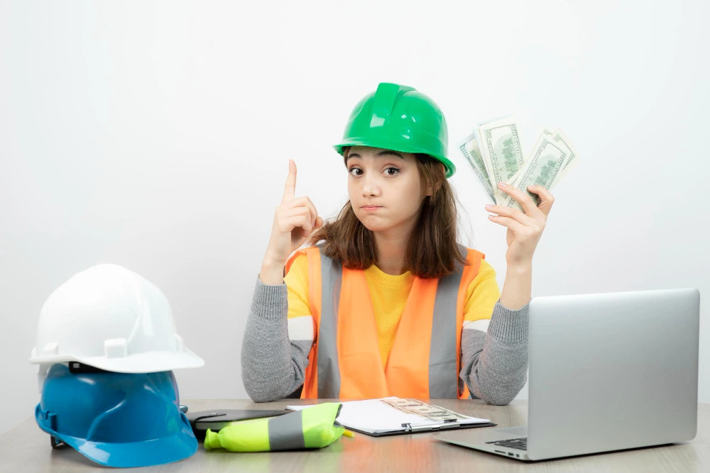 Female construction worker in green hard hat and safety vest holding cash money while pointing up, sitting at desk with laptop and construction helmets