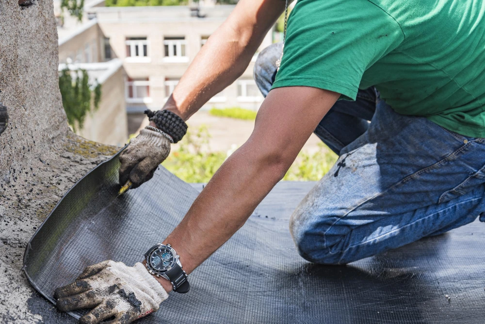 Construction worker in green shirt and work gloves applying waterproofing material to concrete surface using trowel