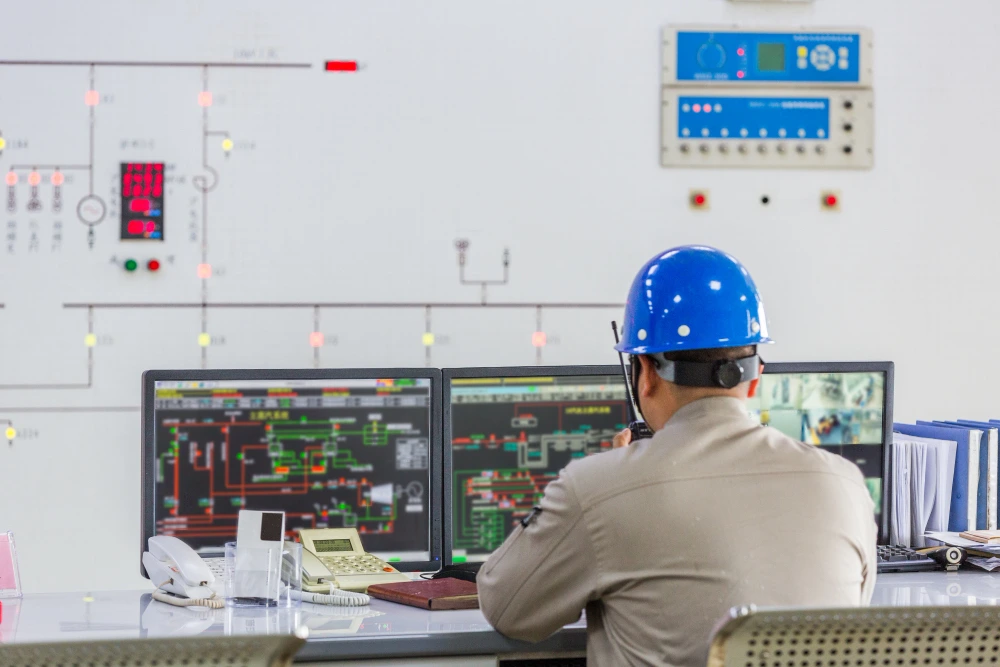 Industrial control room operator in blue hard hat monitoring dual computer screens displaying process control systems and technical diagrams
