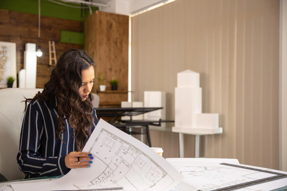Woman in striped shirt examining architectural blueprints at white desk in contemporary office with exposed brick walls and modern lighting