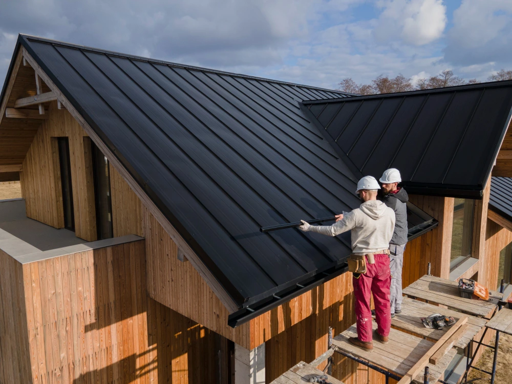 Two construction workers in hard hats installing dark metal roofing panels on a modern wooden house under construction