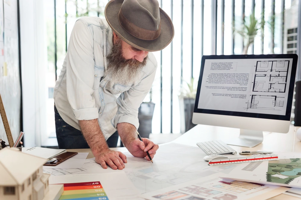 Bearded man in hat and light shirt sketching on papers at desk with computer monitor displaying floor plans, surrounded by design materials and color samples-Restaurant Renovation Estimation Services