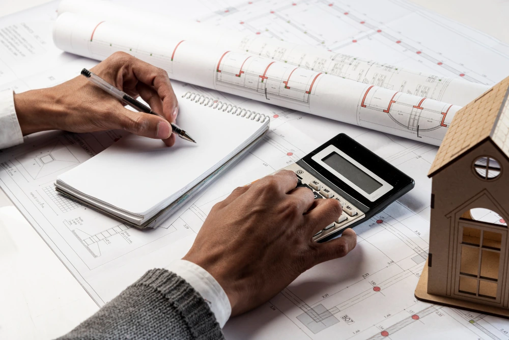 Person in business attire calculating construction costs with calculator and pen over architectural drawings, with rolled blueprints and wooden house model nearby- How We Controlled Costs & Risks on a Multi-Building Project
