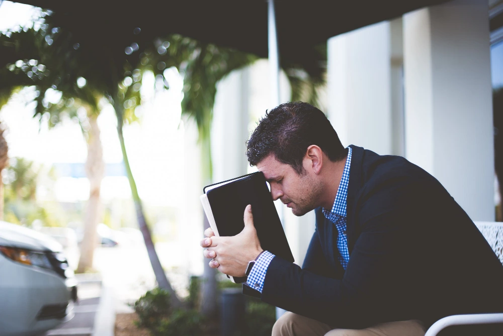 "Stressed businessman in suit holding tablet to his forehead while sitting outdoors, appearing overwhelmed or frustrated