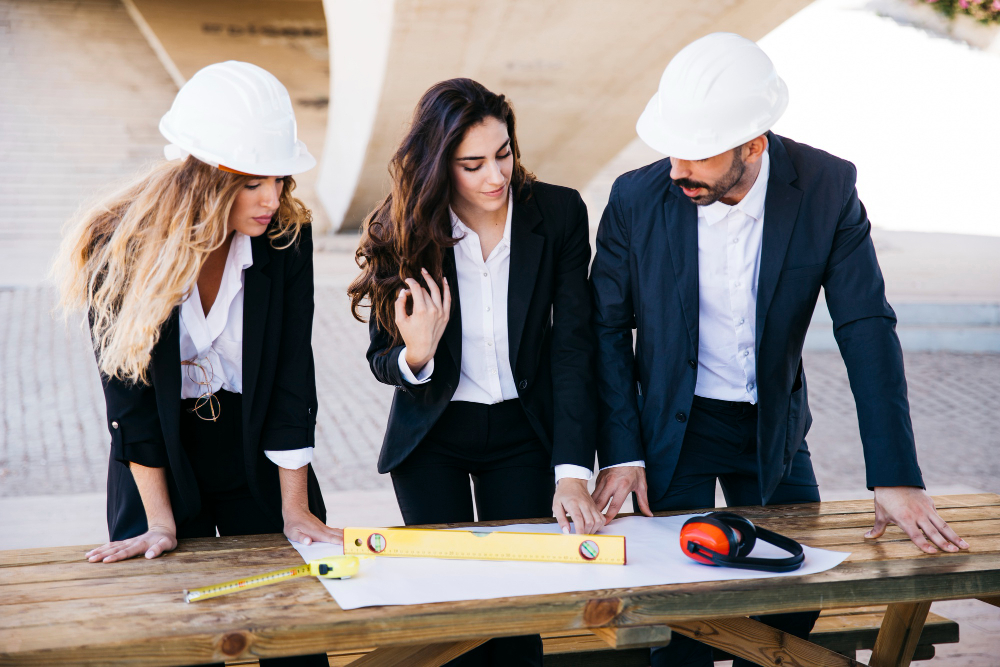 Three construction professionals in business attire and safety helmets examining blueprints and using measuring tools on a wooden table at a construction site