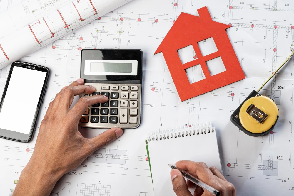 human hands using calculator for construction cost estimation with architectural blueprints, red paper house cutout, smartphone, measuring tape, and notepad on desk