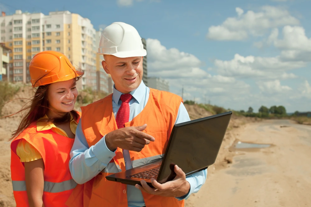 Two construction professionals in hard hats and safety vests reviewing building plans on a laptop computer at an active construction site with apartment buildings in the background