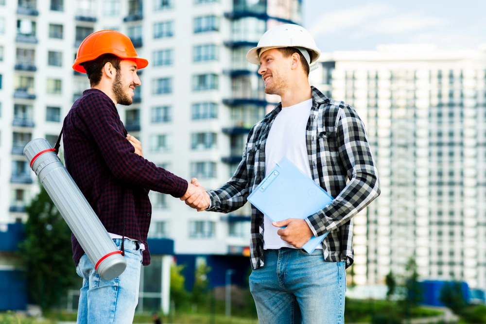 Two male construction workers in hard hats shaking hands outdoors, one wearing orange hard hat with rolled blueprints, other in white hard hat holding blue folder, modern apartment buildings in background- Win More Bids with Accurate Construction Estimation Services