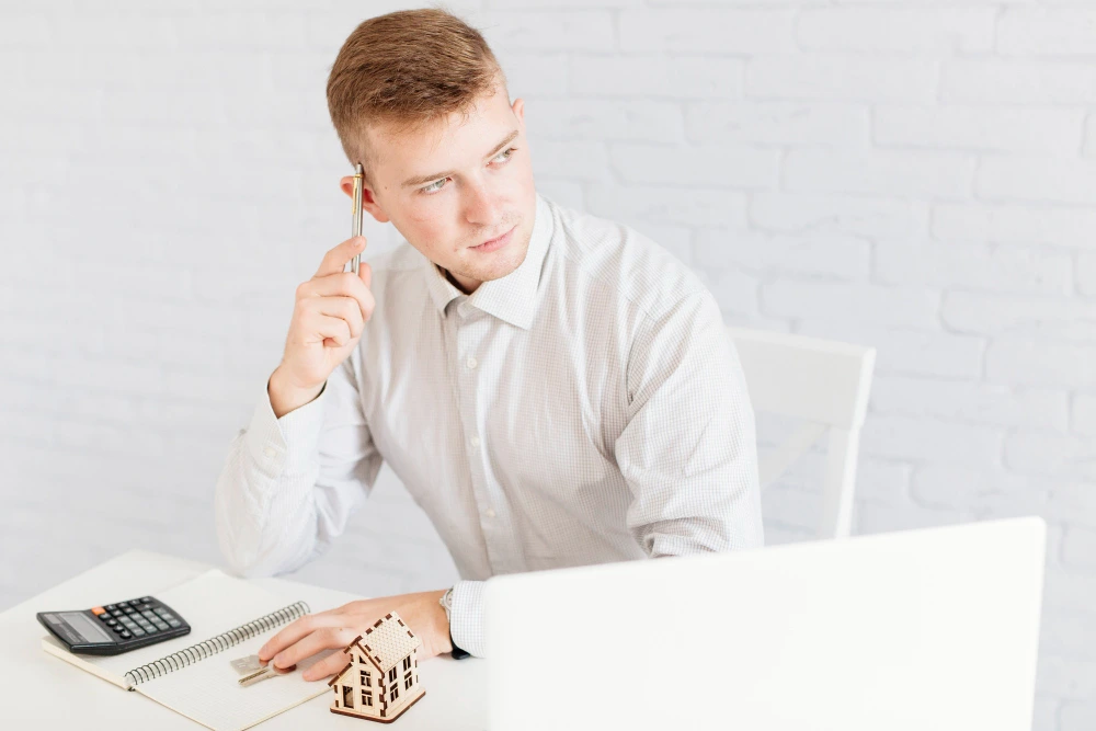 Professional construction estimator in white shirt thinking while reviewing project plans with calculator, notebook, and miniature house model on desk-  Hidden Secrets of Construction Cost Estimation
