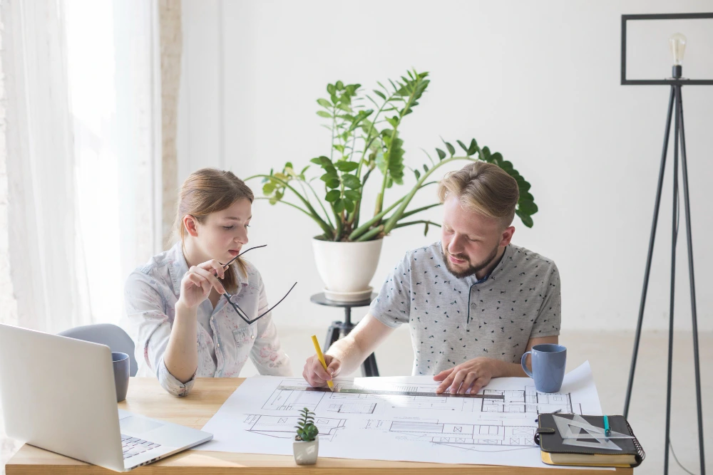 Two construction estimators analyzing architectural blueprints and floor plans on desk with laptop, discussing construction estimation terms and project costs