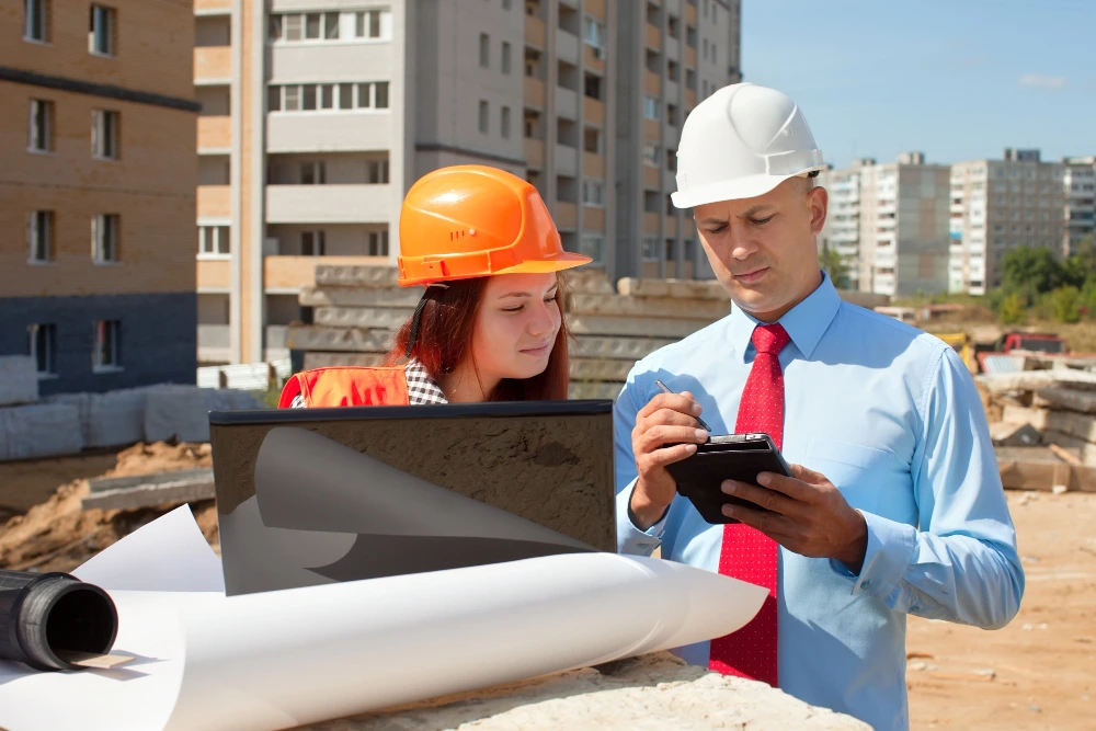 Construction manager and engineer reviewing digital tablet for project estimation at building site with blueprints