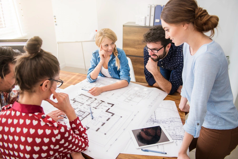A group of designers architects owner and estimaors reviewing blueprints and floor plans on table in casual office setting with tablet nearby