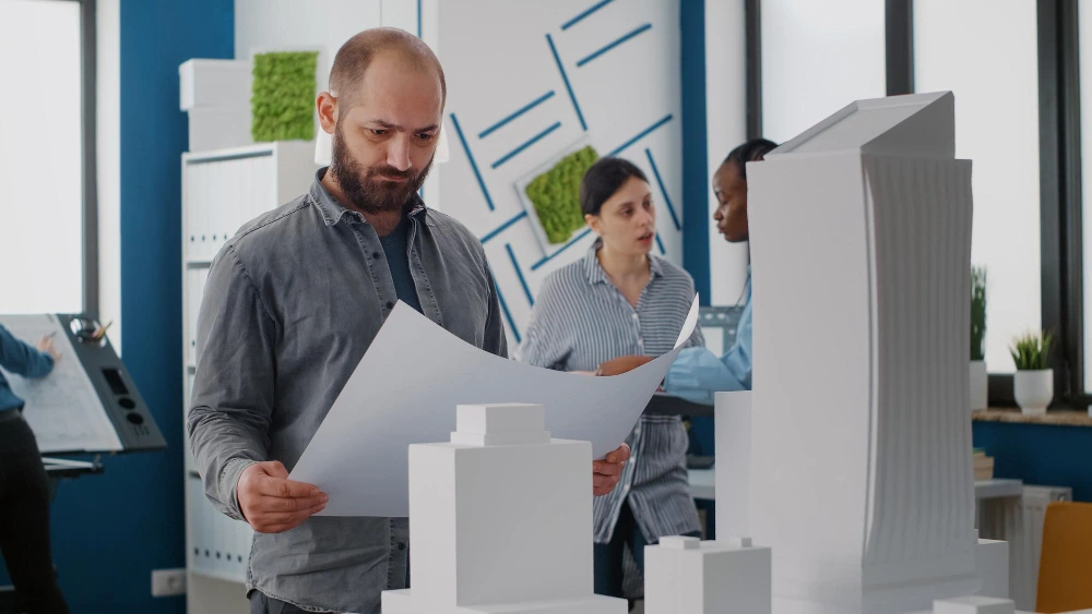 Male estimation expert examining curve architectural drawings while standing next to white curve building model with colleagues discussing about accurate estimation background