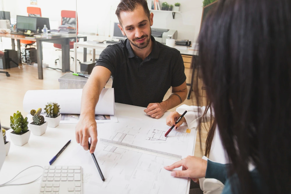 Male construction estimation in black polo shirt discussing architectural shops plans and blueprints with female client at desk in contemporary office workspace- Accurate Shop Fitout Estimation