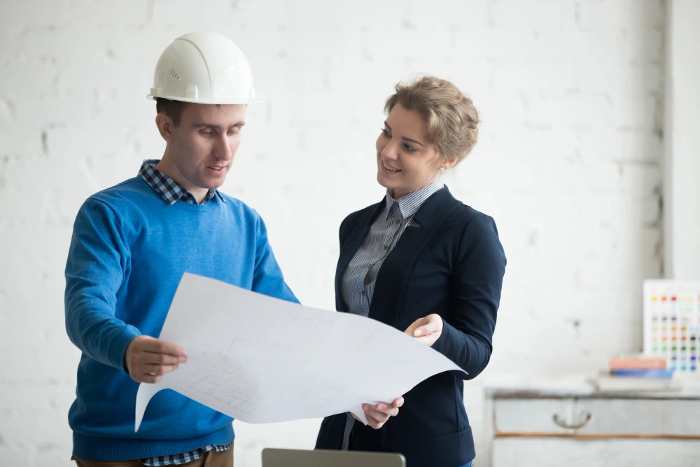 Professional estimation expert in hard hat discussing construction blueprints with female owner during pre-construction planning consultation