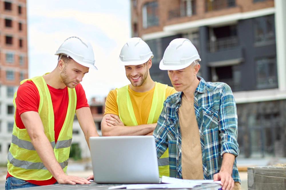3 construction workers in safety gear reviewing laptop for digital construction estimation at commercial building project- New Techniques Are Changing Construction Estimation