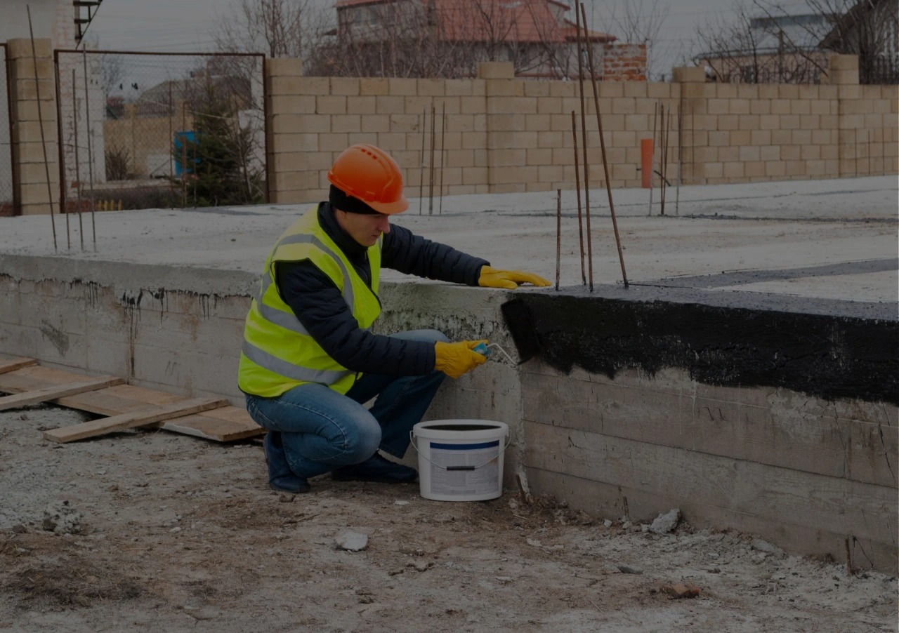 Construction worker in orange hard hat and high-visibility vest applying waterproofing coating to concrete foundation with brush and bucket at building site