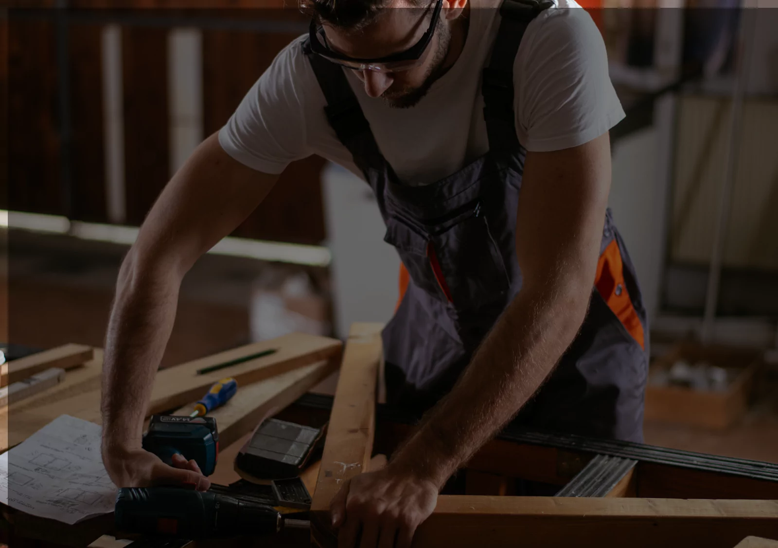 A skilled worker in a t-shirt and overalls using power tools to assemble wooden structures in a workshop