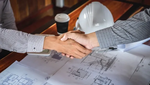 Two people in business attire shaking hands over architectural blueprints and construction plans, with a white hard hat and coffee cup visible on the table